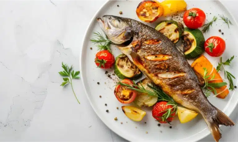 Grilled whole fish with roasted vegetables and herbs on a white plate.