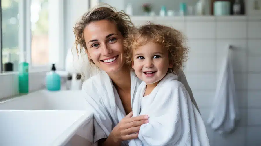 Smiling mother and child in white bathrobes standing together in a bright bathroom.