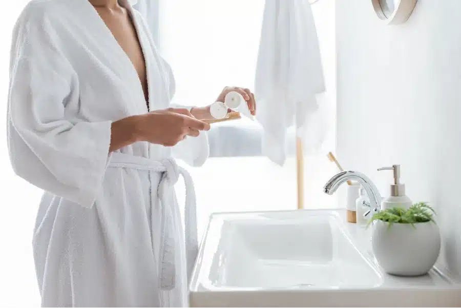 Person in a white bathrobe applying toothpaste to a toothbrush in a modern bathroom.