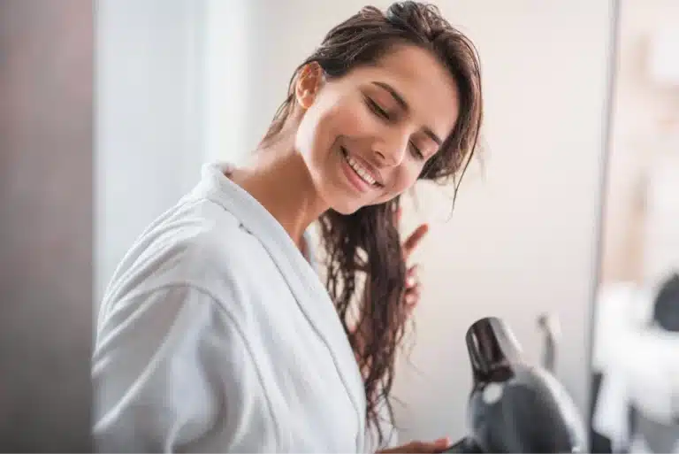Smiling woman drying her wet hair with a black hair dryer, wearing a white bathrobe.