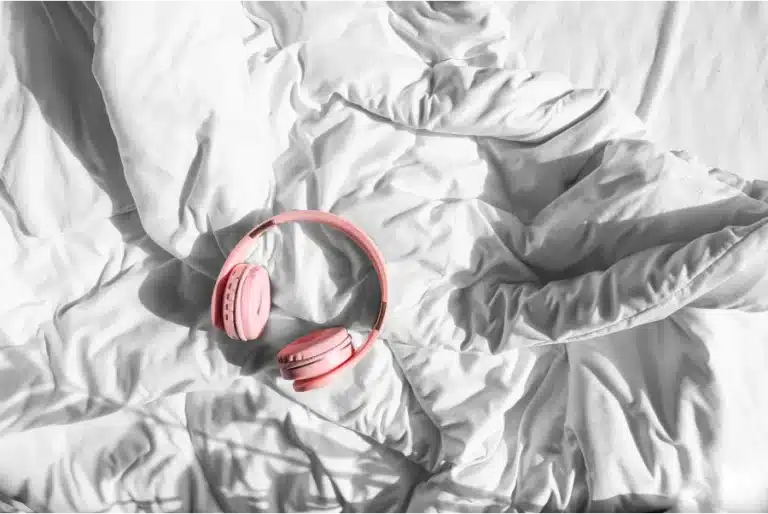 Pink wireless headphones lying on white crumpled bedsheets in soft natural light.