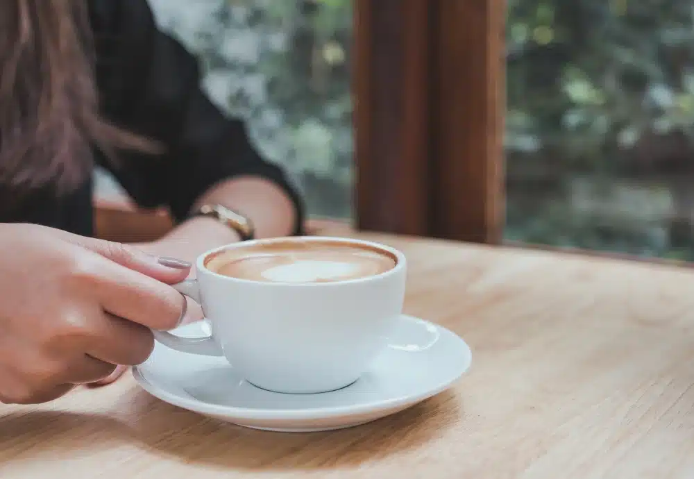 Hand holding a white cup of latte on a wooden table with a blurred window background.