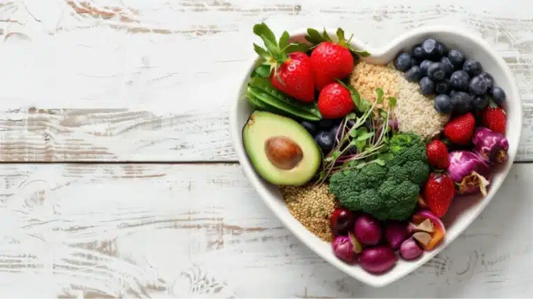 Heart-shaped dish filled with colorful fruits, vegetables, and grains on a white wooden table.