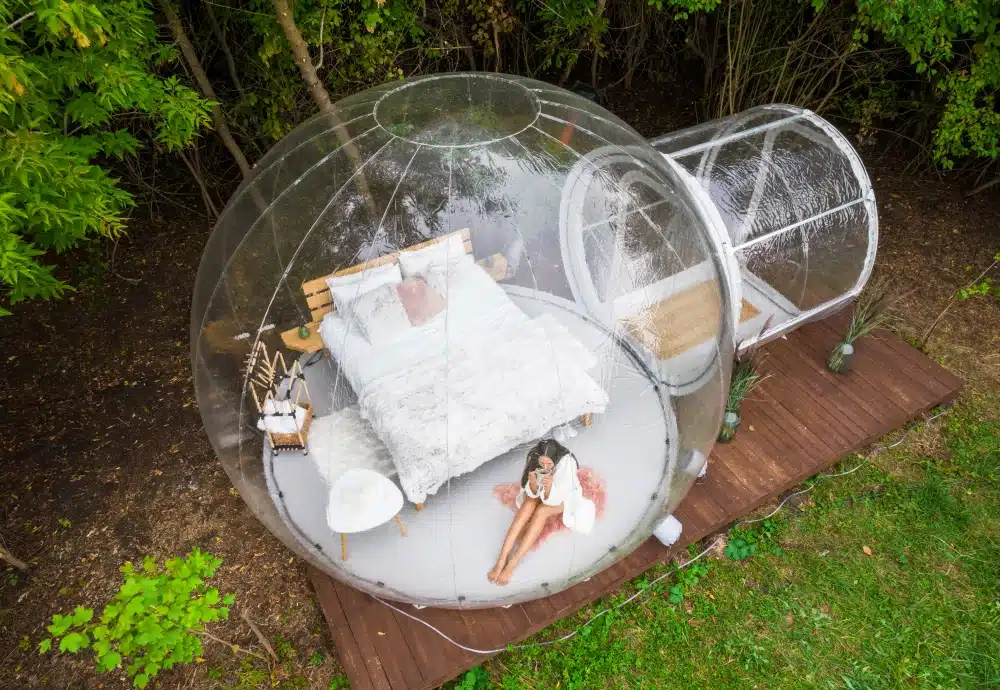Transparent bubble tent with a cozy bed and furnishings set in a forested area.