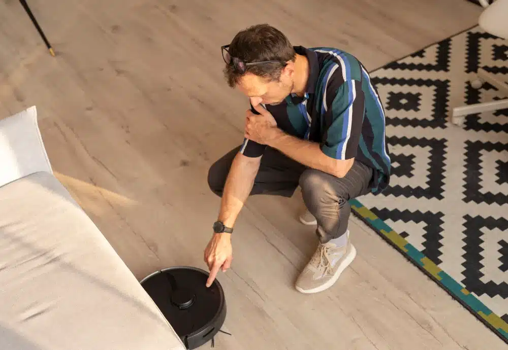 Man kneeling and pointing at a robotic vacuum cleaner on a wood floor.