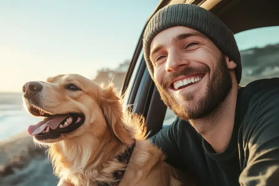 Smiling man with a beanie and golden retriever leaning out of a car window at the beach.